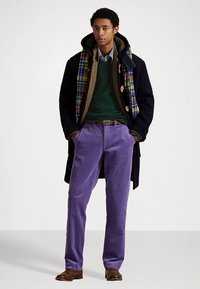 Črni volneni plašč, zelena pulover, vijolične kord hlače, karo šal, rjavi usnjeni čevlji. Priložnostna večplastna obleka z različnimi teksturami in barvami.