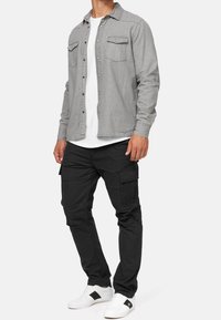 Man in een lichtgrijze overhemd met knopen over een wit T-shirt, zwarte cargobroek en witte sneakers met een zwarte streep op een witte achtergrond.