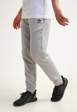 Osoba ubrana w jasnoszare spodnie dresowe Adidas i czarne buty sportowe Adidas, stojąca z lekko uniesioną jedną stopą na białym tle.