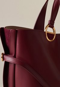 Burgundy leather handbag featuring dual textures, smooth and pebbled. Gold hardware ring accent and structured silhouette with stitching detail.