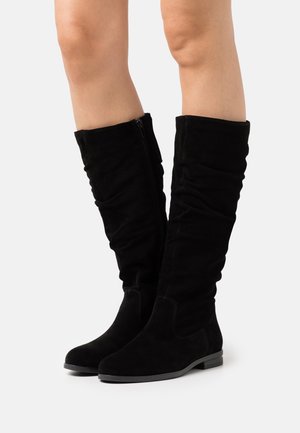 Black suede knee-high boots with a slouched design, side zipper, flat sole, and minimal stitching details. Smooth texture and slim profile.