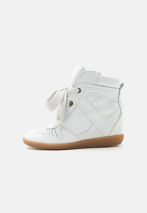 Sneakers alte bianche con spesse stringhe bianche, area della punta perforata, collo imbottito e suola in gomma naturale su sfondo semplice.