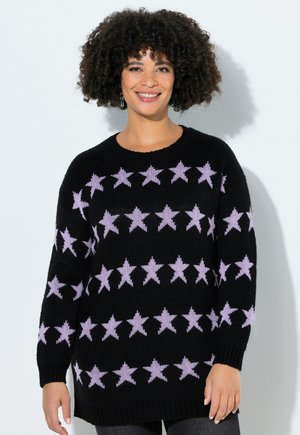Smilende kvinde med krøllet hår iført sort sweater med mønster af vandrette rækker af pink stjerner, stående mod hvid baggrund.