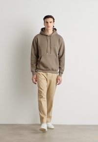 Sudadera marrón con cordones, bolsillo frontal, pantalones beiges y zapatillas blancas; de pie sobre un fondo neutro.