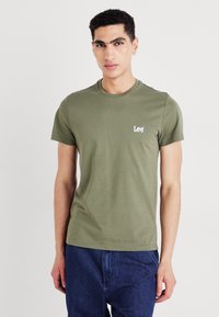 T-shirt en coton vert olive avec un col rond, des manches courtes et un petit logo "Lee" blanc sur la poitrine gauche. Design simple et coupe ajustée.