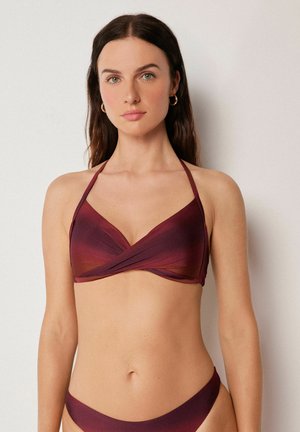 Frau mit langen braunen Haaren, die einen burgunderfarbenen, verlaufenden Neckholder-Bikini und goldene Creolen vor einem schlichten Hintergrund trägt.