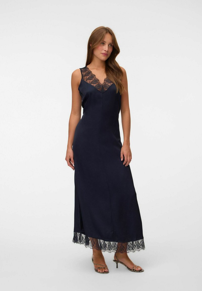 Femme portant une robe maxi noire sans manches avec un bord en dentelle au niveau du décolleté et de l'ourlet, associée à des sandales à talons ouvertes.