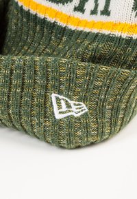 Bonnet en tricot vert avec une texture côtelée, des rayures jaune et blanche, et un écusson logo brodé blanc sur le revers.