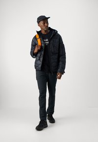 Chaqueta de plumas acolchada negra con capucha, combinada con una camiseta gráfica negra y pantalones oscuros. Correas naranjas en la mochila negra. Zapatillas negras.