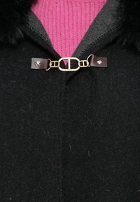 Cappotto di lana nera con colletto in pelliccia sintetica, dotato di una chiusura con gancio oro e marrone. Sotto c'è un pullover lavorato a maglia rosa con una texture particolare.