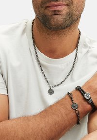 Zwarte metalen ketting met een kompas hanger, gecombineerd met een gevlochten leren armband met een ronde kompasdetail, gedragen tegen een witte shirt.