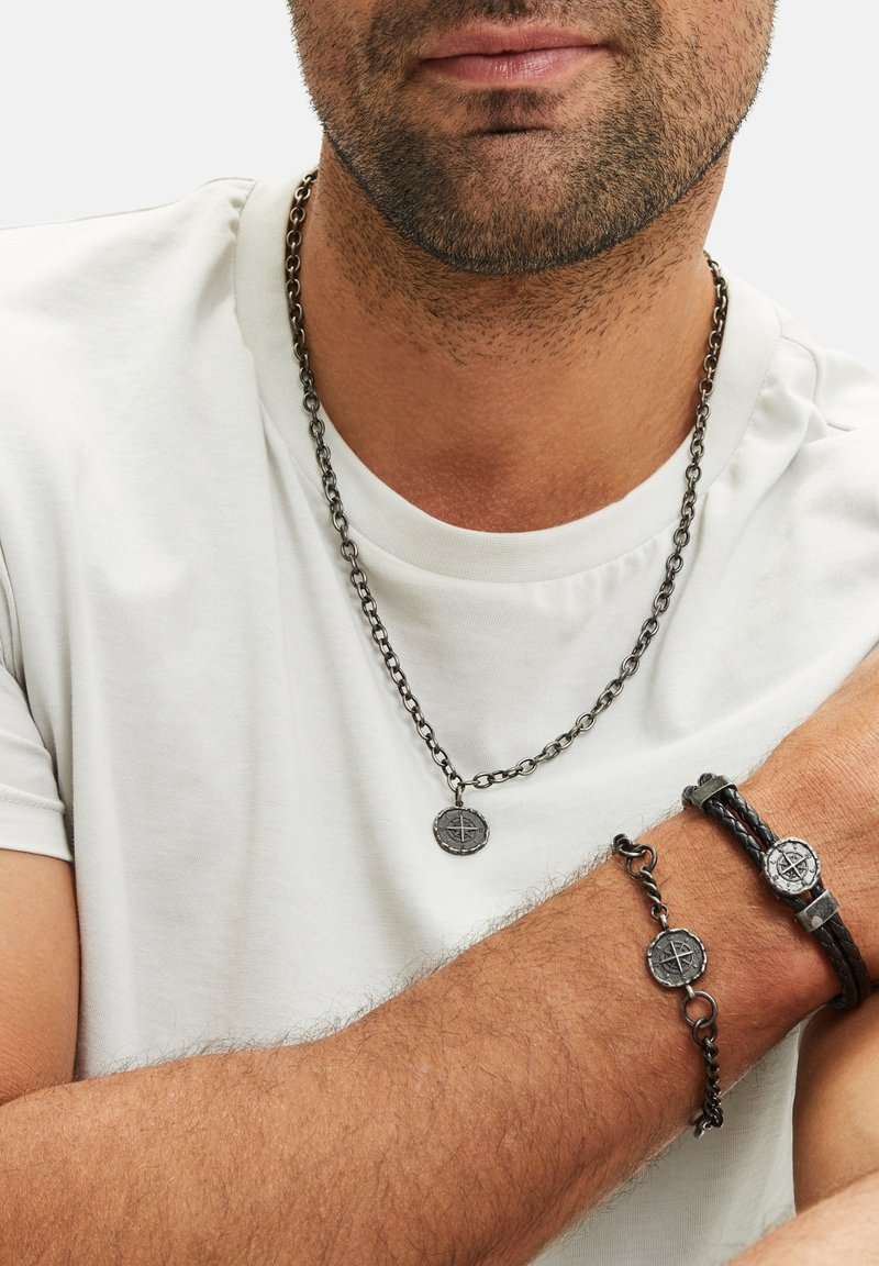 Zwarte metalen ketting met een kompas hanger, gecombineerd met een gevlochten leren armband met een ronde kompasdetail, gedragen tegen een witte shirt.
