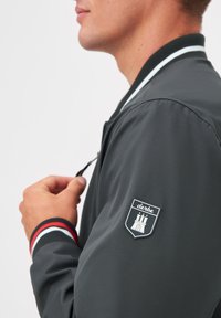 Grå bomberjacka med svart krage och randiga ärmslut. Har en badge på vänster ärm. Slät textur, dragkedjestängning och slim fit-design.