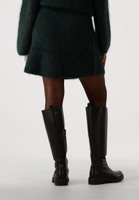 Kvinne iført gjennomsiktige svarte strømpebukser, et mørkegrønt ullskjørt og genser, samt høye svarte skinnstøvler som står foran en hvit bakgrunn.