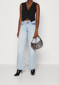 Kvinna i svart ärmlös kort topp, ljusblå högmidjade vida byxor, svarta spetsiga stövlar, håller i en handväska med leopardmönster.