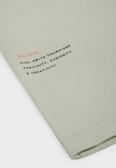 Tela de algodón verde claro con textura de acanalado que presenta texto impreso en rosa y negro. El texto dice: “Paul Smith defiende la positividad, la curiosidad y la creatividad.”