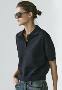 Marineblauer kurzärmeliger Polopullover aus strukturiertem Strick. Mit flachem Kragen und einer Knopfleiste mit drei Knöpfen, kombiniert mit blauen Jeans.