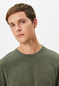 Grön bomullströja med rund halsringning och texturerad yta, ribbad krage och avslappnad passform, visad mot en neutral bakgrund.