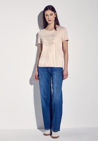 T-shirt pêche à manches courtes avec un design textuel en encre dorée, associée à un jean large bleu et des baskets blanches, se tenant devant un fond pâle.