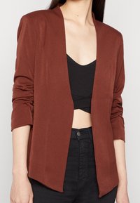 Blazer v měděné barvě s elegantním designem, otevřeným předním dílem a vyhrnutými rukávy, vyrobený z měkké tkaniny; nošený přes černý zkrácený top.