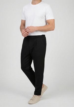 Uomo in piedi che indossa una maglietta bianca a maniche corte semplice, pantaloni neri e sneaker beige con lacci, su uno sfondo neutro.