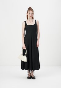 Melnā bezpiedurkņu maksi kleita ar kvadrātveida dekoltē, gluda tekstūra, kombinēta ar baltu rokassomu un melnām šņorējamām kurpēm. Minimalistisks dizains.