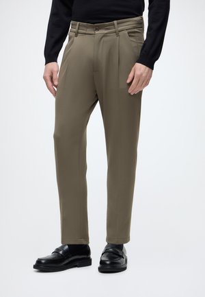 Hombre vestido con pantalones de vestir verde oliva, suéter negro y mocasines de cuero negro, de pie sobre un fondo claro y sencillo.