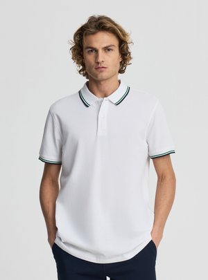 Giovane uomo con capelli ricci che indossa una polo bianca con bordature a righe verdi e nere, in piedi con le mani nelle tasche su uno sfondo semplice.