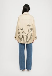 Giacca oversized beige con stampa floreale nera sul retro, dotata di colletto e dettagli con bottoni, abbinata a pantaloni in denim larghi blu chiaro.