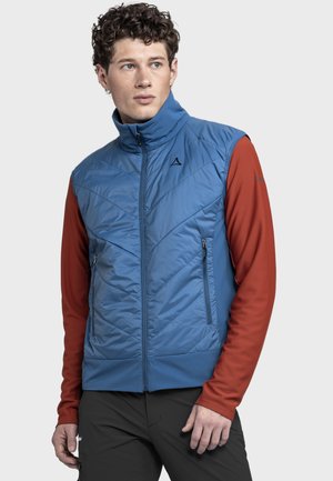 Junger Mann mit lockigem Haar, der eine blaue gesteppte Weste über einem langärmligen roten Hemd und schwarzen Hosen trägt, schaut zur Seite.