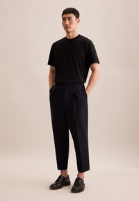 T-shirt nera a maniche corte abbinata a pantaloni a pieghe blu navy, figura in piedi con le mani nelle tasche e scarpe nere lucide.