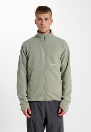 Junger Mann trägt eine hellgrüne Fleecejacke mit Reißverschluss und dunkelgraue Hose, steht vor einem schlichten weißen Hintergrund.