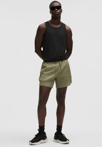 Melna bez piedurknēm tvertnes blūze ar zaļiem teksturētiem šortiem, pieguļošs jostasvieta un melni sporta apavi. Vīrietis valkā tumšus saulesbrilles.