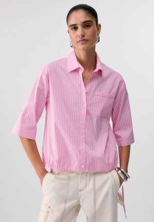 Donna con capelli pettinati all'indietro che indossa orecchini a cerchio d'argento, una camicia a righe rosa e bianche con bottoni e pantaloni beige chiaro con le mani in tasca.