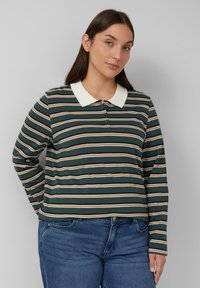 Junge Frau mit langen dunklen Haaren, die ein gestreiftes Langarm-Polohemd in Grün, Beige und Weiß sowie eine blaue Jeans trägt, steht vor einem grauen Hintergrund.