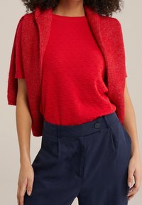 Röd texturerad tröja bärs med en röd kortärmad top, i kombination med marinblå högmidjade byxor som har en enkel knappdetalj.