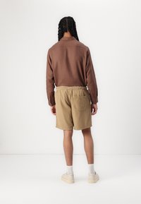 Chemise marron à manches longues avec col, associée à un short en velours côtelé beige. Le short possède une taille élastique et une poche arrière. Chaussettes et chaussures blanches.