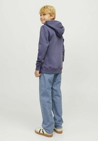 Paarse hoodie met een kanga pocket, gecombineerd met lichtblauwe jeans. De hoodie heeft een trekkoord en ribbelmanchetten. Witte schoenen met groene accenten.