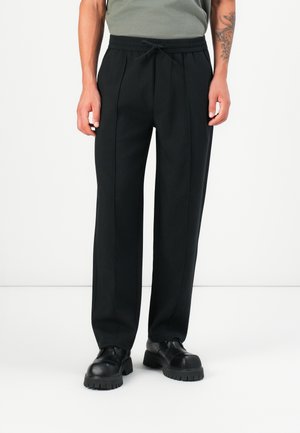 Schwarze Hosen mit einem lockeren Schnitt, ausgestattet mit einem elastischen Bund und einem Kordelzug. Glattes Material, schmal zulaufende Beine und dezente Frontfalten.
