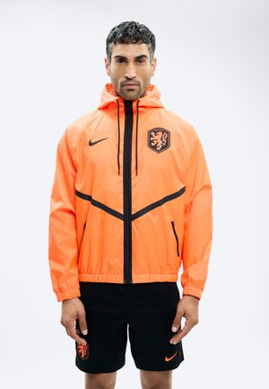 Man draagt een feloranje sportjack met capuchon, voorzien van een zwart leeuwembleem, en zwarte korte broek met bijpassende logo's, staand tegen een effen achtergrond.
