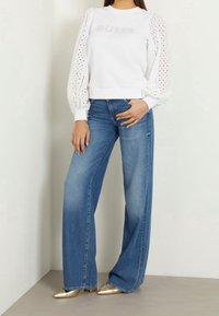 Sudadera blanca con mangas abullonadas perforadas y logo "GUESS". Combinada con jeans azules de pierna ancha y zapatos dorados de punta fina.