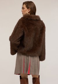 Donna con una giacca in finta pelliccia marrone, una gonna a quadri con pieghe rosse, orecchini a cerchio dorati e stivali in pelle marrone alti fino al ginocchio, di profilo.