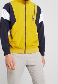 Chaqueta bloqueada en color amarillo y azul marino con detalles blancos, cierre de cremallera y logo en el pecho. Presenta puños elásticos y bolsillos.