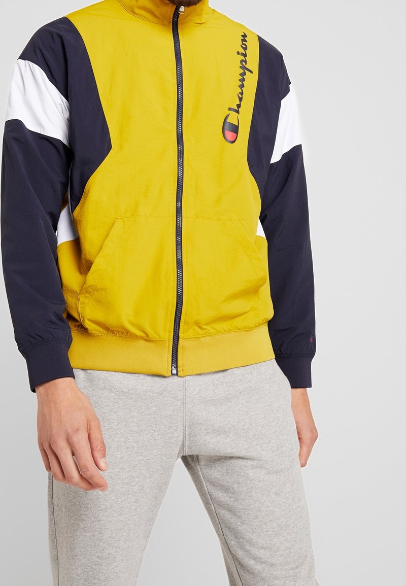 Chaqueta bloqueada en color amarillo y azul marino con detalles blancos, cierre de cremallera y logo en el pecho. Presenta puños elásticos y bolsillos.