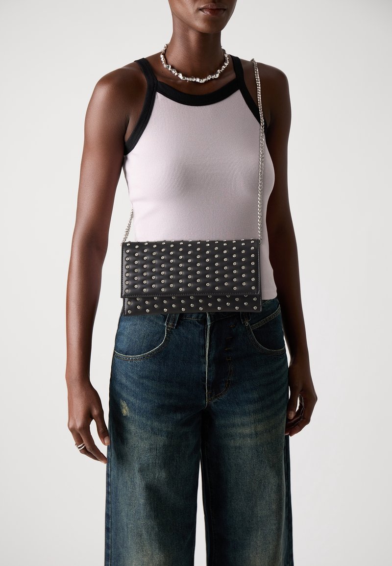 Femme portant un top blanc et noir sans manches, un jean bleu à jambes larges, un collier argenté et un sac à épaule noir clouté avec une bandoulière chaîne.