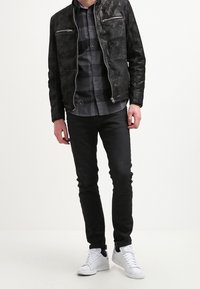 Schwarze Jacke aus Kunstleder mit zwei Reißverschlusstaschen, über einem dunklen karierten Hemd getragen, kombiniert mit schwarzer Jeans und weißen Turnschuhen.