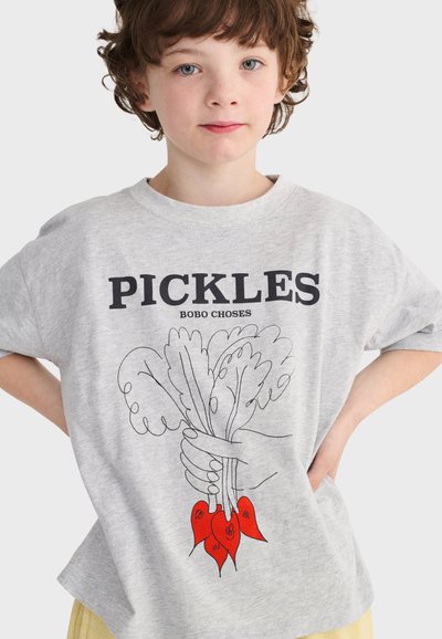 Kind mit kurzen lockigen Haaren, das ein graues T-Shirt trägt und eine Hand zeigt, die grüne Blätter mit roten Wurzeln hält, mit dem Text "PICKLES BOBO CHOSES".