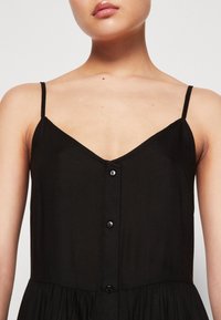 Žena v černých šatech bez rukávů s tenkými ramínky a předními knoflíky, odhalující krk, ramena a horní část hrudníku.