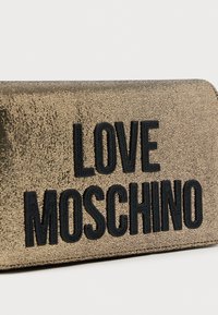 Πορτοφόλι με χρυσαφένια υφή και ρωγμές, με ανάγλυφα μαύρα γράμματα "LOVE MOSCHINO." Ορθογώνιο σχήμα με κλείσιμο με φερμουάρ.