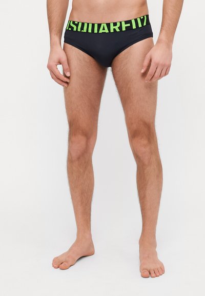 Modello maschile che indossa slip da bagno neri con una larga fascia in vita verde e nera con la scritta "SQUARE".